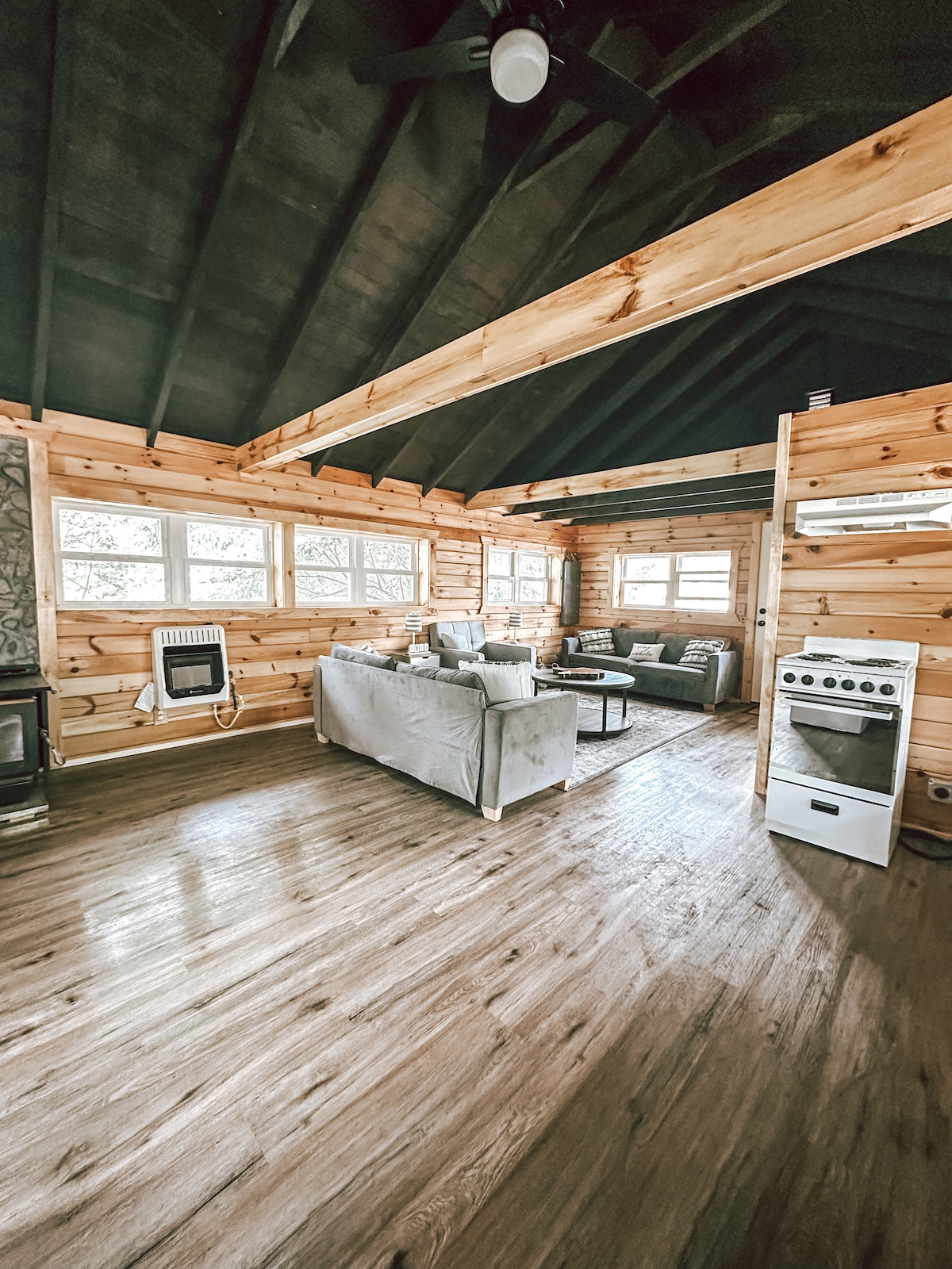 A warm living area is highlighted by wood paneling and a high ceiling. A comfortable sofa and armchair are arranged around a central coffee table. Natural light filters through multiple windows, with a vintage stove positioned near the wall, adding a touch of charm.