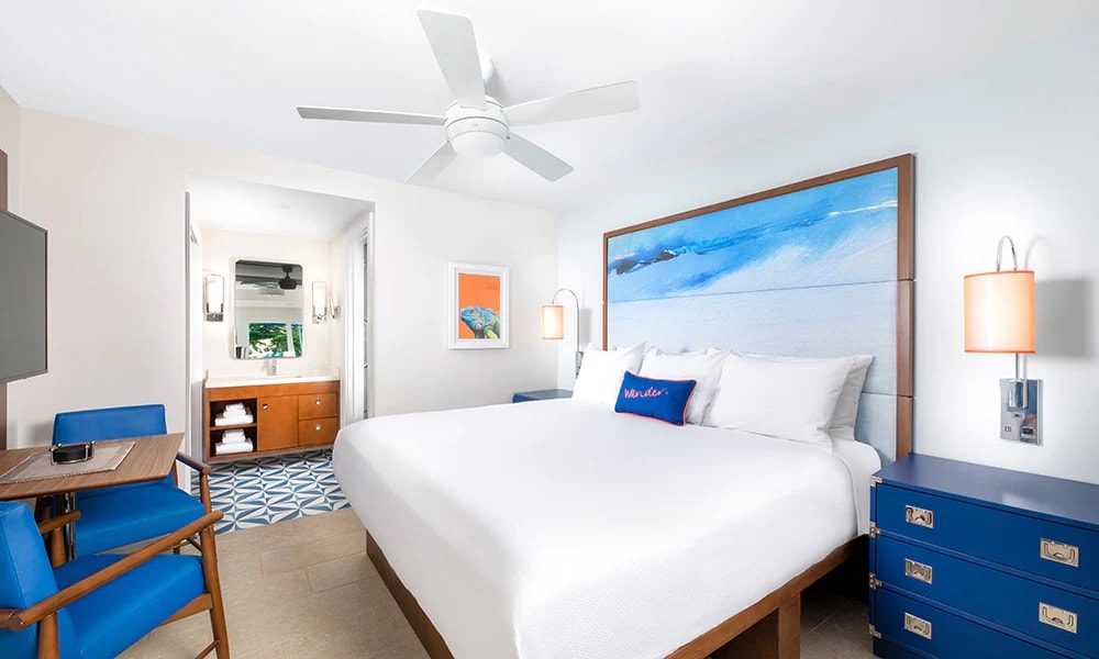 A spacious sleeping area features a large bed with white linens and a vibrant headboard depicting a serene blue landscape. A ceiling fan circulates air above, while a wooden desk and chair complement blue storage units. A doorway leads to a separate area with additional furnishings.