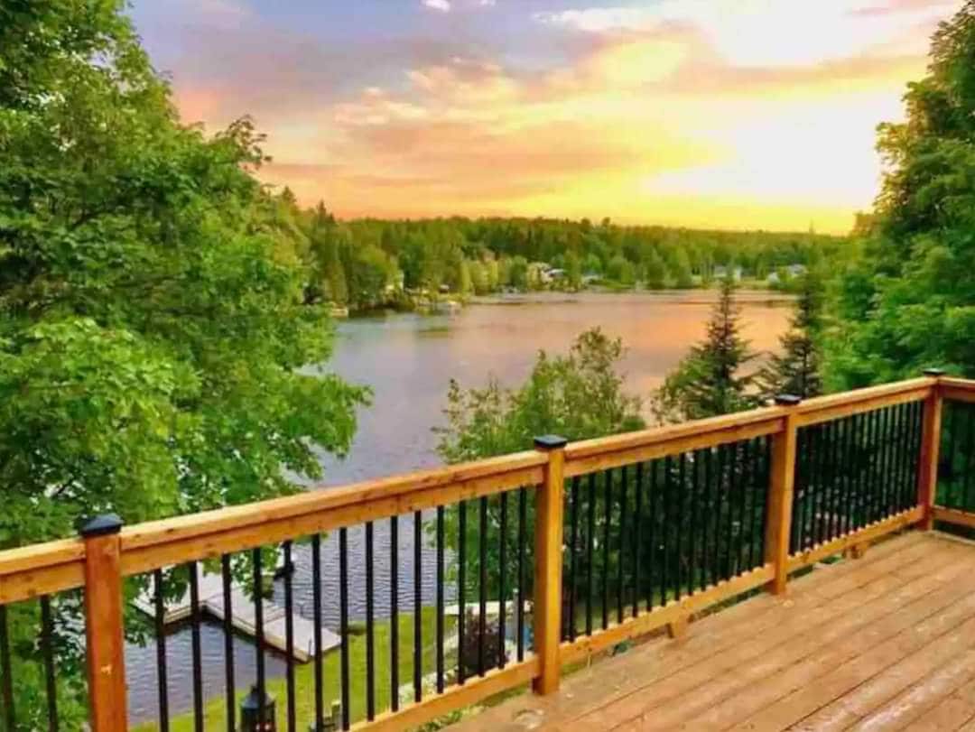 A wooden deck overlooks a serene lake, framed by lush greenery. The calm water reflects the vibrant colors of a sunset, enhancing the peaceful setting. The scene is complemented by distant trees and a glimpse of a dock along the lake's edge.