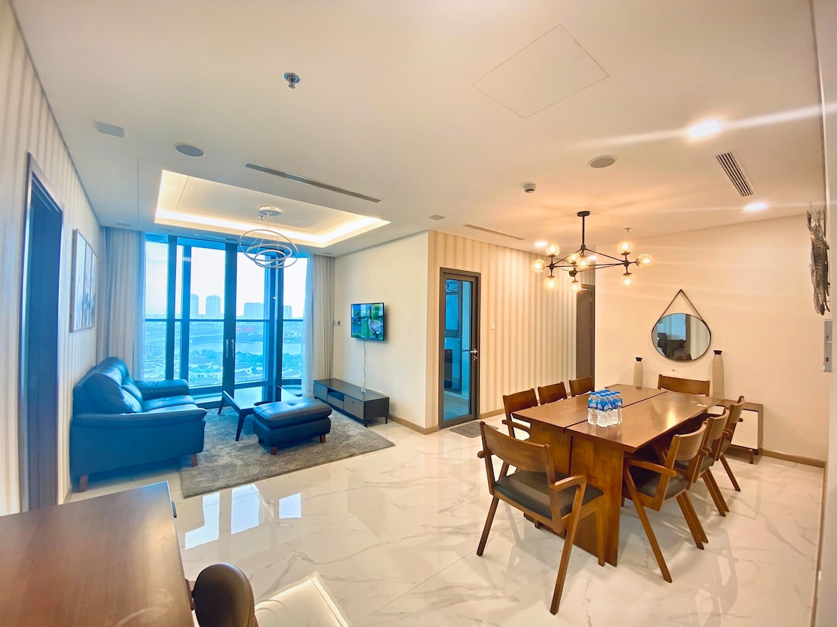 A spacious living and dining area features sleek furniture and large windows offering city views. A modern sofa and ottoman sit beside a wall-mounted television. A wooden dining table is surrounded by six chairs, with a decorative mirror and light fixture enhancing the contemporary design.