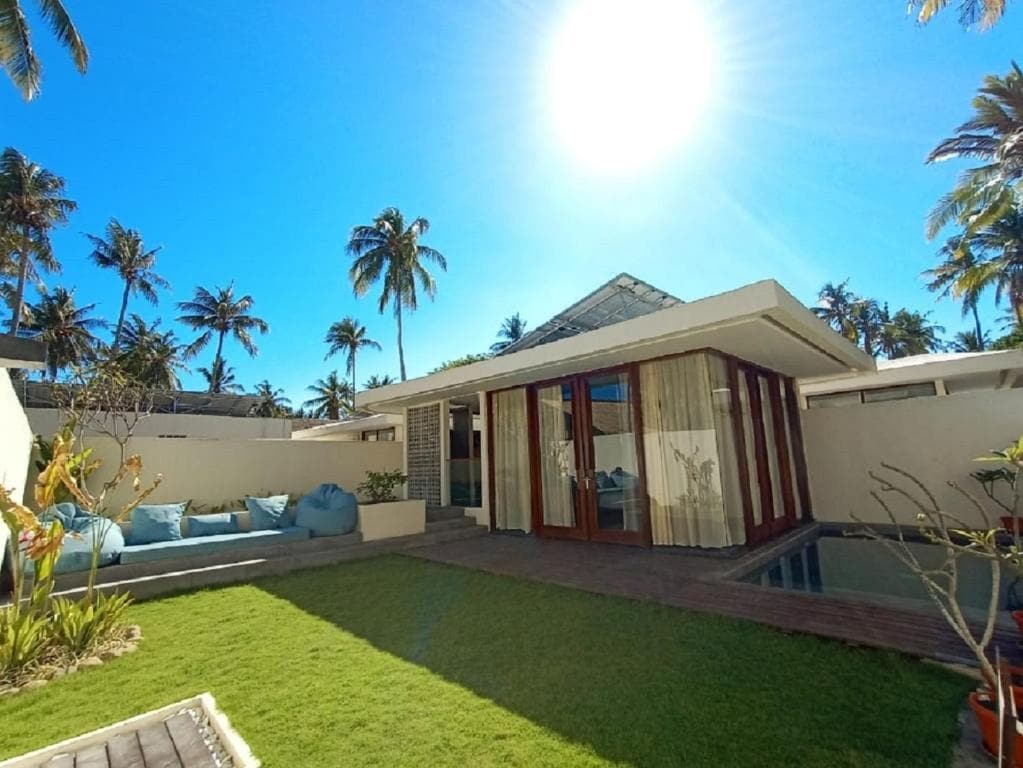 A modern villa is surrounded by a lush garden with palm trees and vibrant greenery. Large glass doors open to an inviting living space, and a private plunge pool is visible beside a wooden deck. Sunlight casts a warm glow over the entire scene.