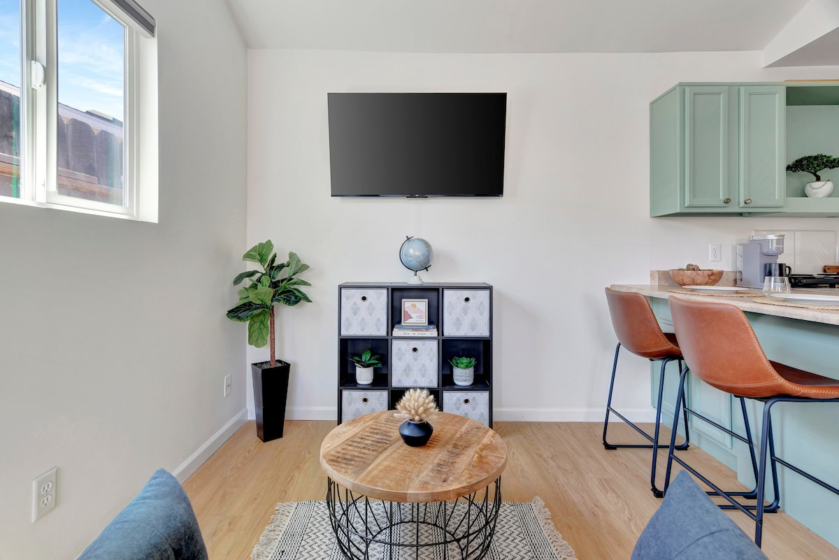 A bright and modern living area features a mounted TV above a stylish storage unit. A small wooden coffee table sits on a textured rug, surrounded by two comfortable chairs. Soft natural light enters through the window, and a green plant adds a touch of nature.