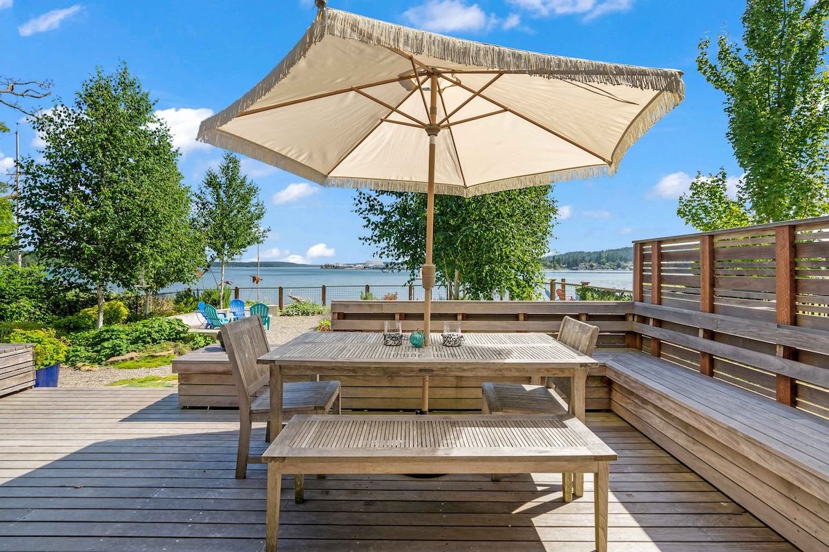 An outdoor deck is presented with a large wooden dining table and benches, shaded by a beige umbrella. Lush greenery surrounds the area, with views of the water in the background, inviting relaxation and outdoor dining.