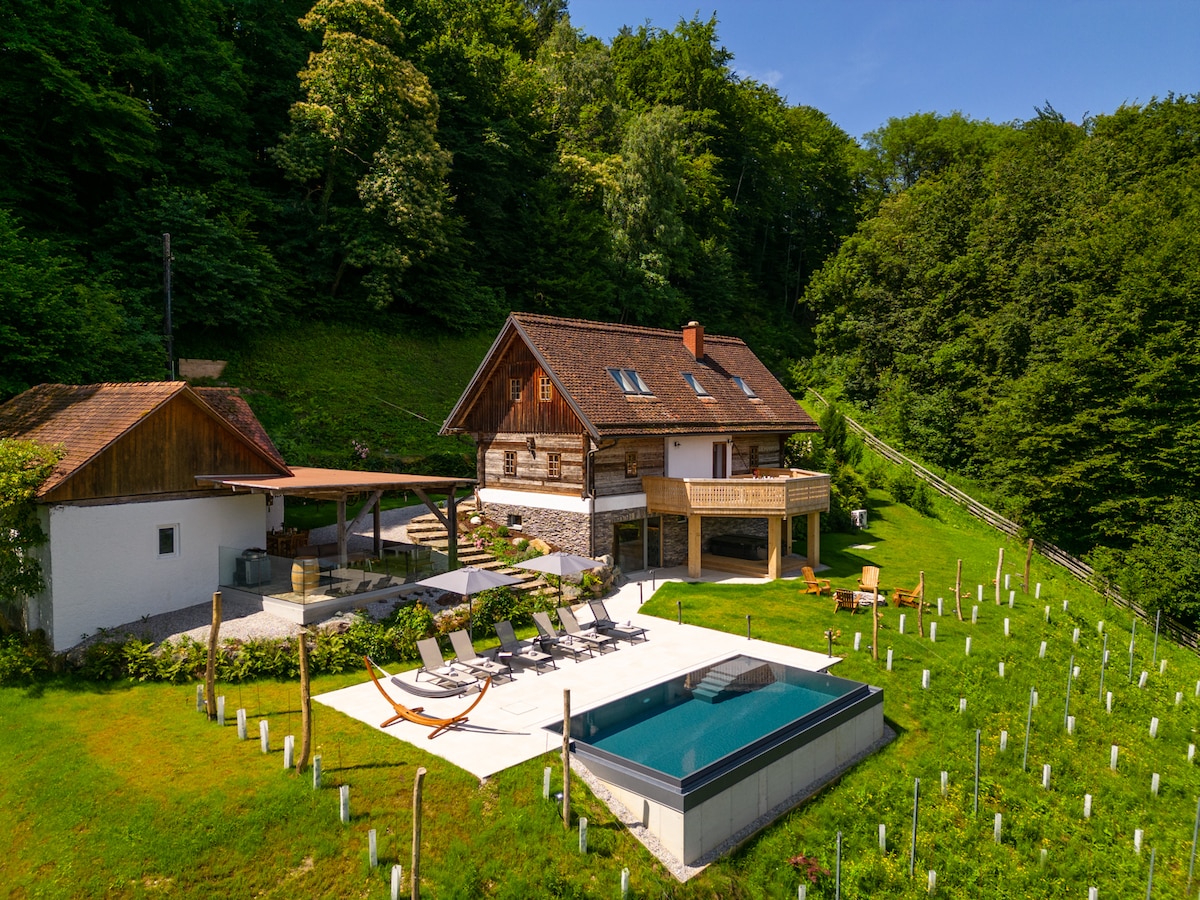 The chalet is set within a lush green landscape, featuring a spacious outdoor area with a heated infinity pool and several sun loungers. The building's wooden facade blends with the surrounding nature, and a terrace extends from the structure, offering a serene escape in a private setting.