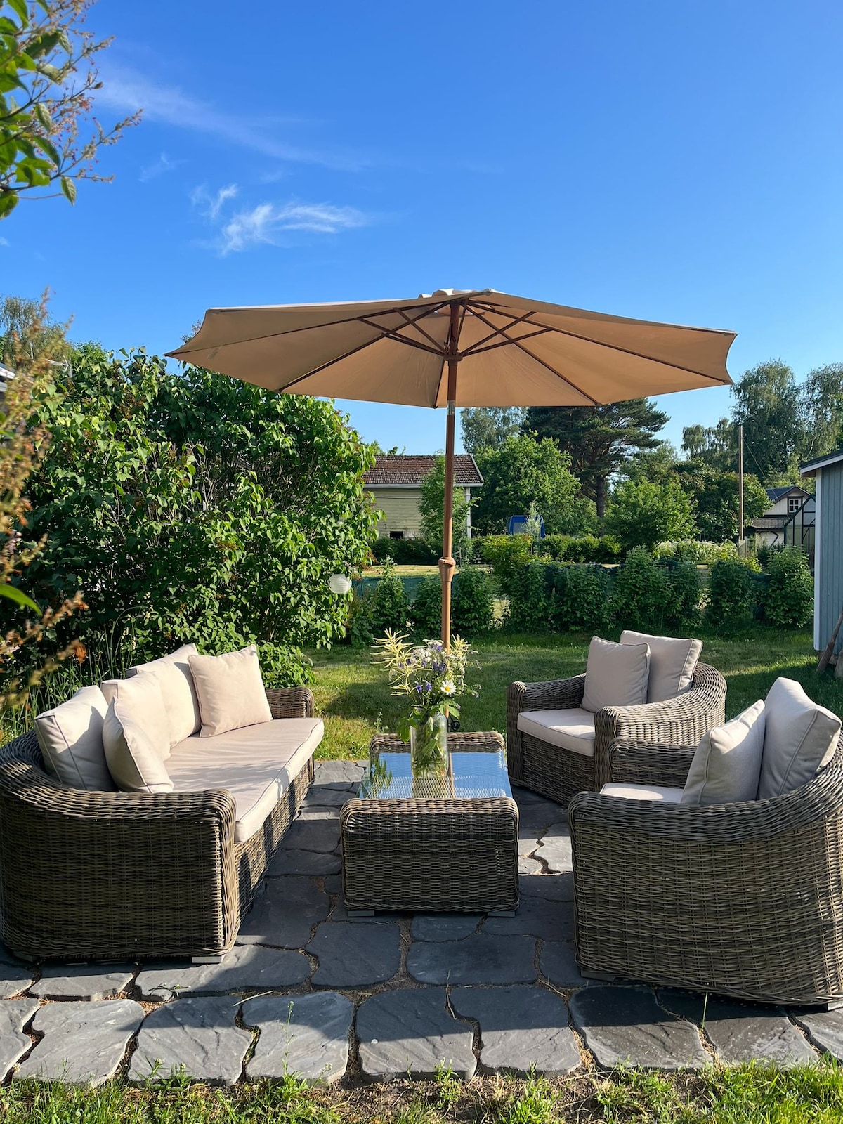 A relaxing outdoor seating area is arranged on a stone patio. Two cushioned armchairs and a connecting loveseat surround a glass-topped table. A large umbrella provides shade, while lush greenery is visible in the background, enhancing the tranquil garden setting.