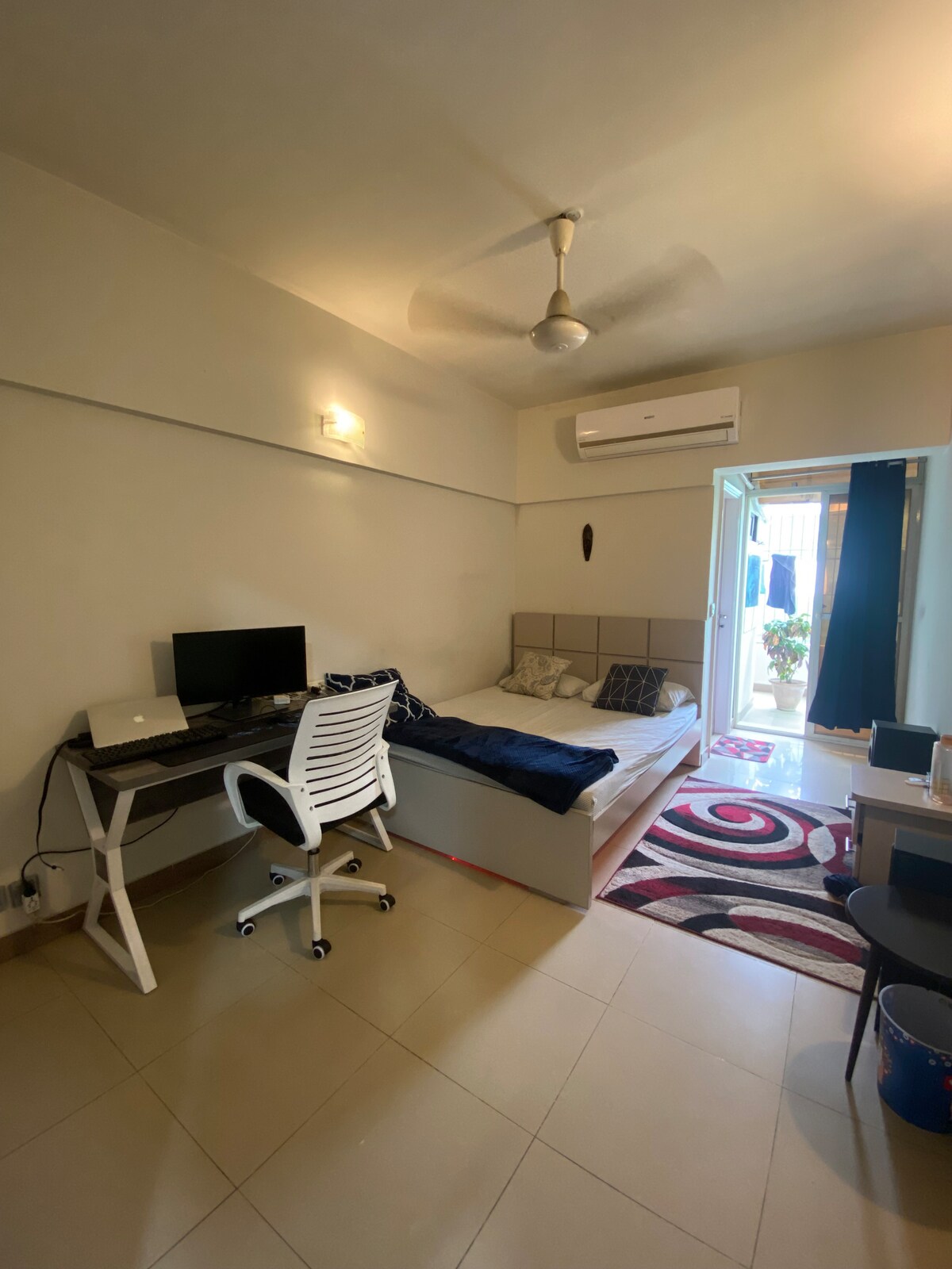 A well-lit room features a spacious bed against the wall, with a desk and chair positioned nearby. A monitor is placed on the desk, and light spills in from the open door, leading to a small outdoor space adorned with greenery.