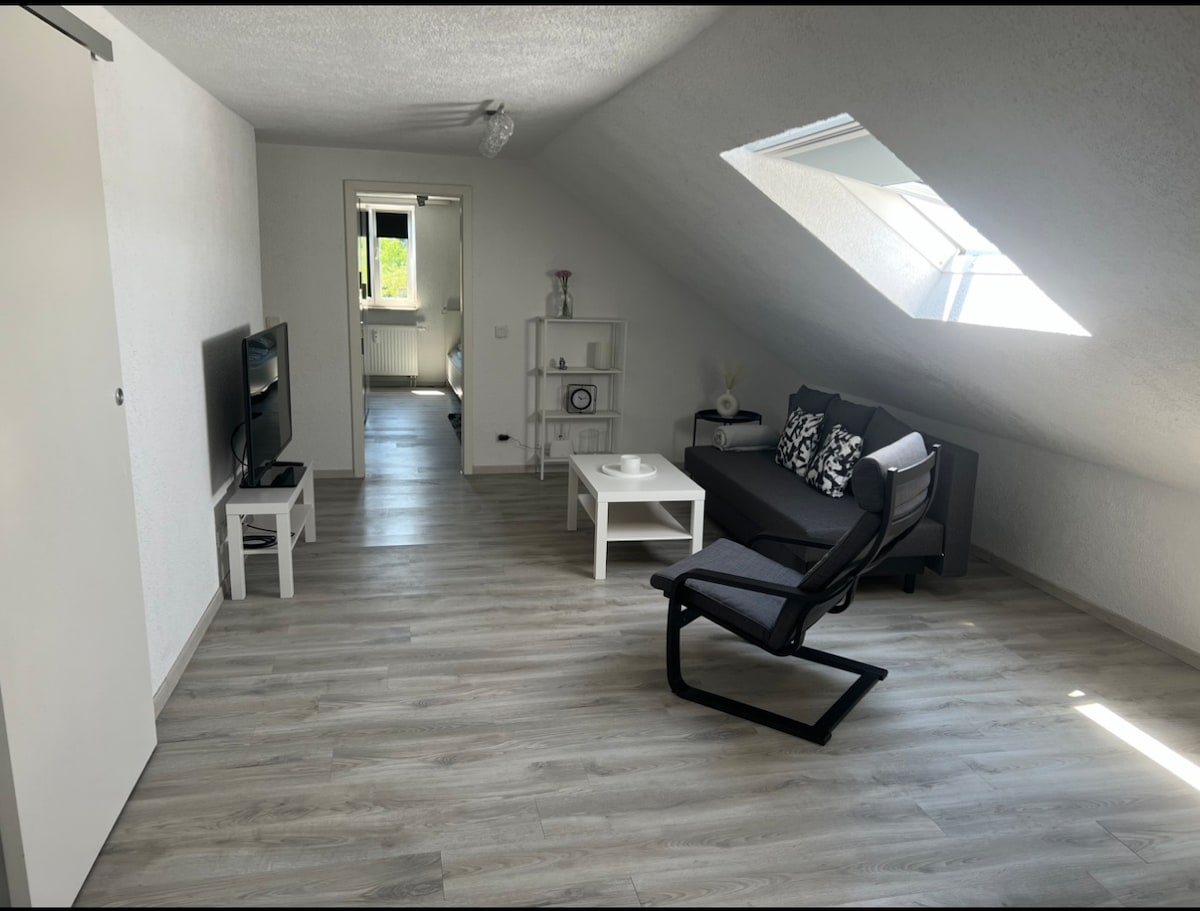 An inviting living area features a comfortable dark sofa, coffee table, and a television. A sleek black chair is positioned nearby. Natural light enters through a skylight, illuminating the space. Light-colored flooring enhances the modern aesthetic of the room.