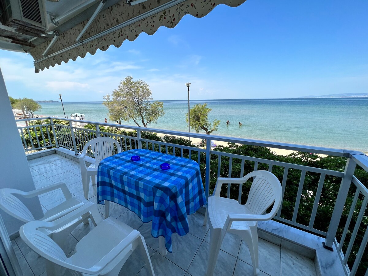 An outdoor terrace is depicted with a checkered blue tablecloth covering a table, surrounded by several white chairs. The coastline with gentle waves can be seen in the background, framed by trees, under a clear blue sky.
