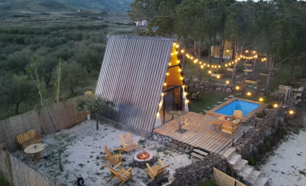 An aerial view captures the unique triangular structure of the bungalow, surrounded by a scenic landscape. Soft lighting decorates the outdoor area, which features a mini pool, a fire pit, and wooden seating. Lush greenery and well-maintained pathways enhance the serene environment.