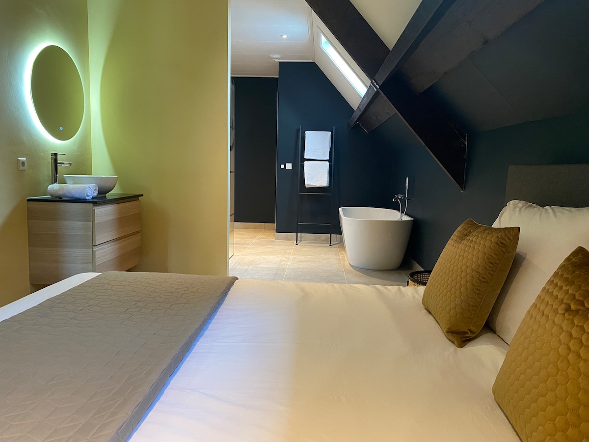 The spacious bedroom features a comfortable bed with two textured pillows. A freestanding bathtub is visible in the background, alongside a modern vanity sink. The room is illuminated by a circular mirror light, with wooden beams enhancing the ceiling.