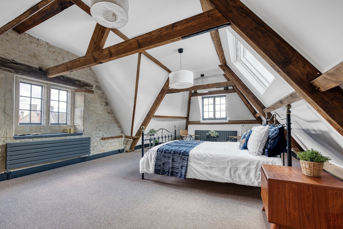 A spacious master bedroom features exposed Cotswold stone walls and distinctive wooden beams. The room is furnished with a large bed, a bedside table, and has two skylights allowing natural light. A comfortable seating area is provided, with a cozy blanket draped over the bed.