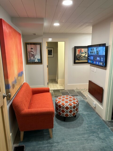 A welcoming hallway features an orange couch and a decorative ottoman with a geometric pattern. Colorful artwork adorns the walls, while a flat-screen TV is mounted nearby. Soft lighting from recessed fixtures illuminates the space, leading to a doorway at the end.