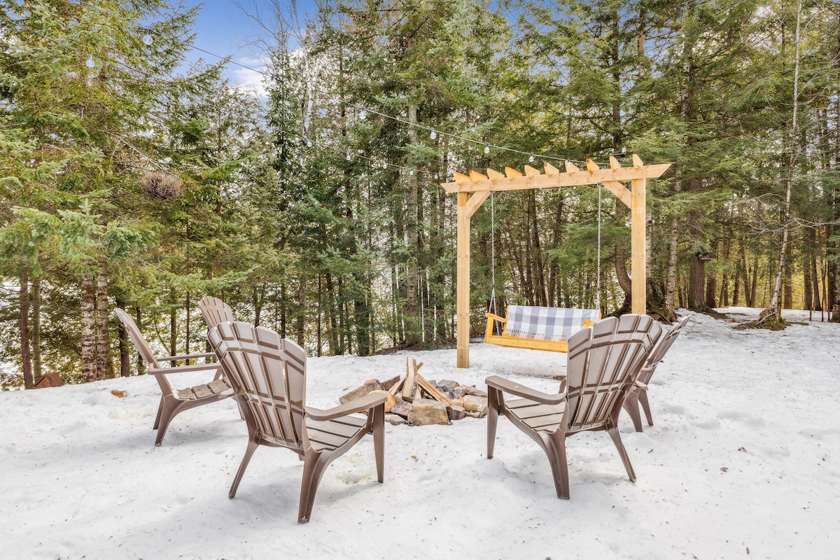 A fire pit surrounded by four outdoor chairs is set in a snowy landscape, with a wooden pergola in the background. A cozy swing is hung near the pergola, and tall evergreen trees provide a natural backdrop.