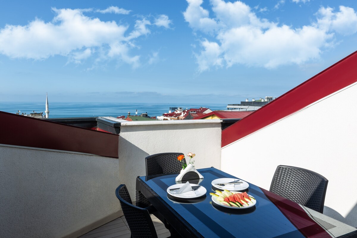 A rooftop terrace displays a dining table set with plates and fresh fruit. Black wicker chairs surround the table, while a small vase with flowers adds a touch of elegance. A panoramic view of the sea and distant rooftops is visible under a bright blue sky.