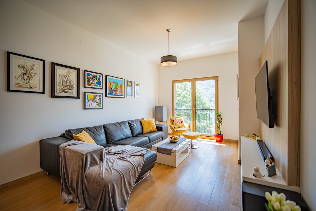 A modern living area features a comfortable sectional sofa adorned with yellow cushions and a throw blanket. Art pieces are displayed on the wall, adding color to the space. A coffee table sits in front, with bright natural light illuminating the room through large windows.