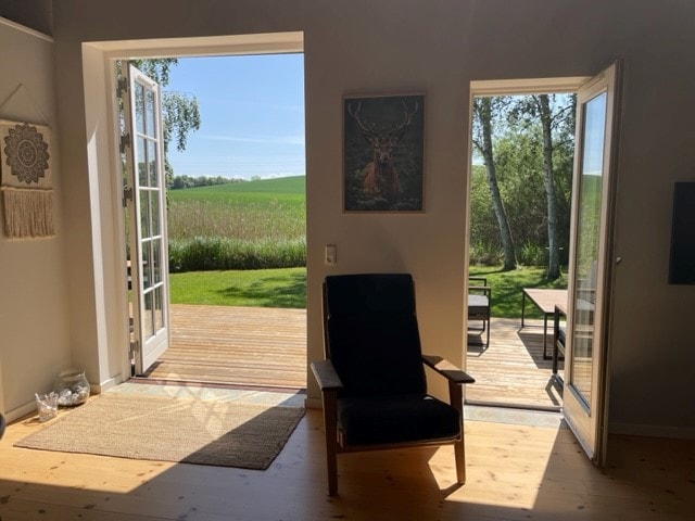 A bright living area is shown with two open French doors leading to a wooden deck. Natural light floods the room, highlighting a black armchair and a textured rug. A serene view of greenery and a distant field is visible outside.