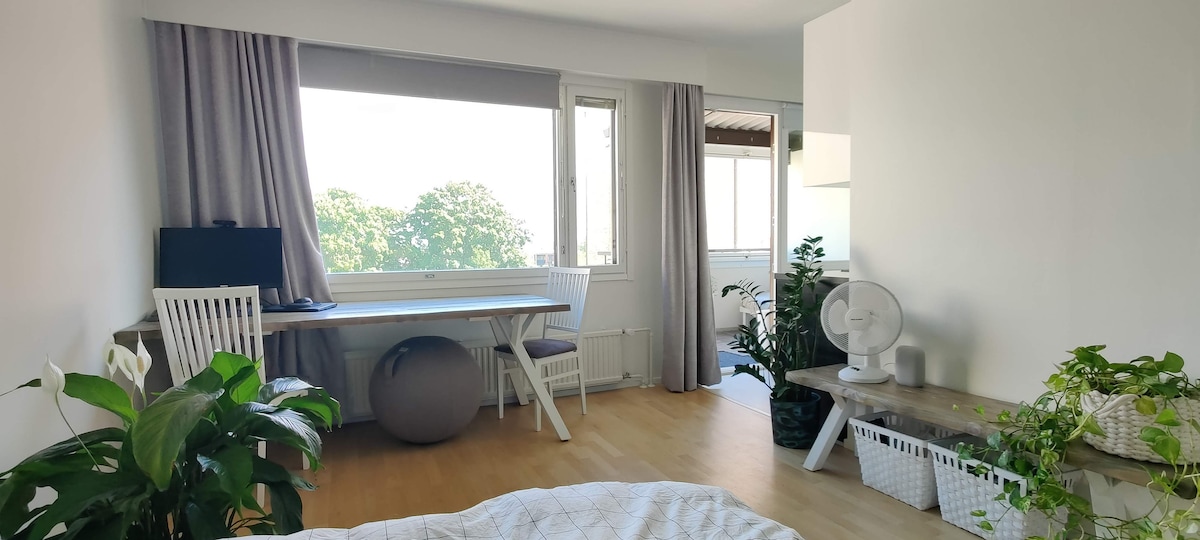 A bright studio space features a workspace with a computer monitor, positioned near a window. Natural light fills the room, which includes a white table and wooden benches. Potted plants line the area, offering a touch of greenery. A fan is placed strategically for comfort.
