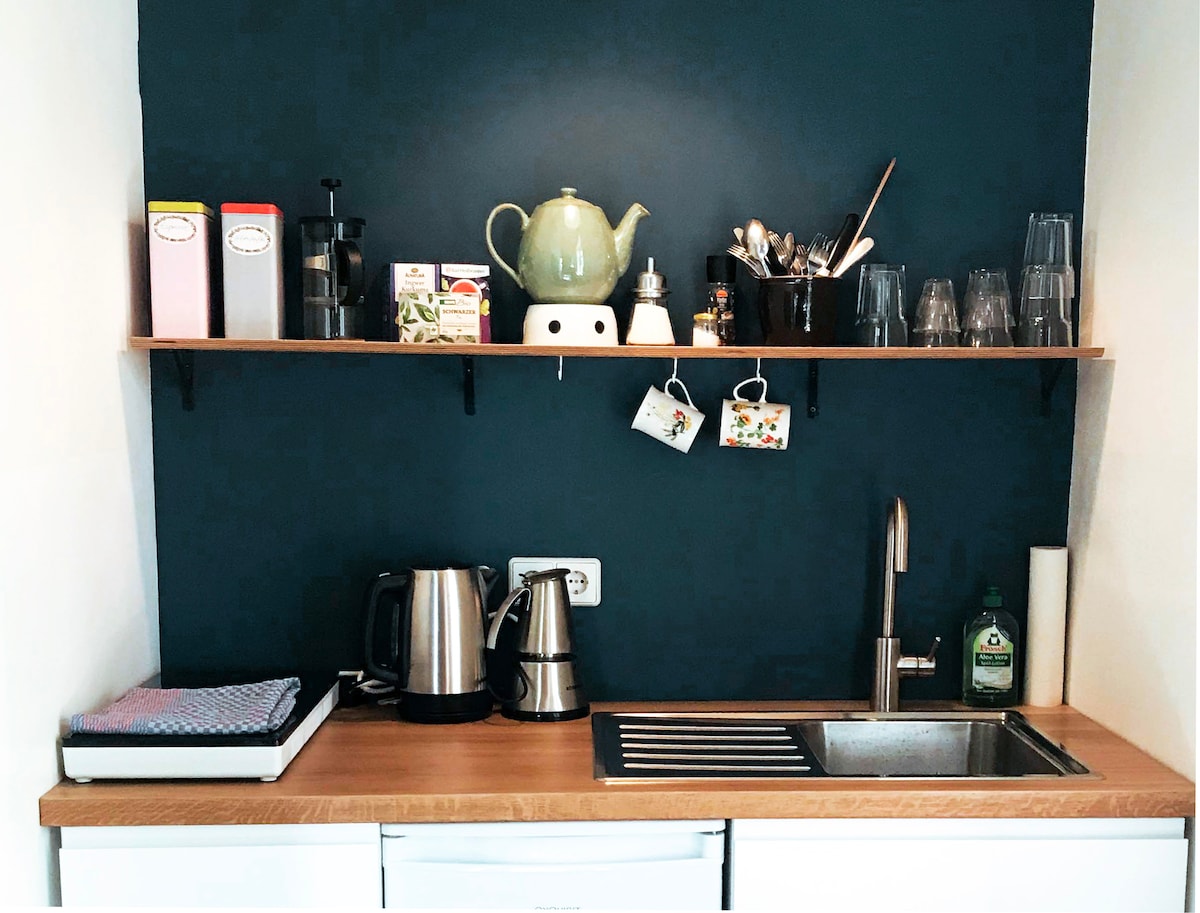 A compact kitchen space features a wooden countertop with a stainless-steel sink. Above the countertop, a shelf holds various utensils, coffee equipment, and glassware. Two electric kettles are placed on the counter along with a dish drying rack, enhancing practicality.