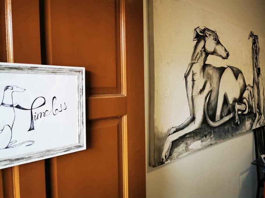 An entryway features a stylish sign reading 'Timeless' next to a large, monochromatic artwork depicting reclining greyhounds. The warm wooden door adds a welcoming touch to the space.