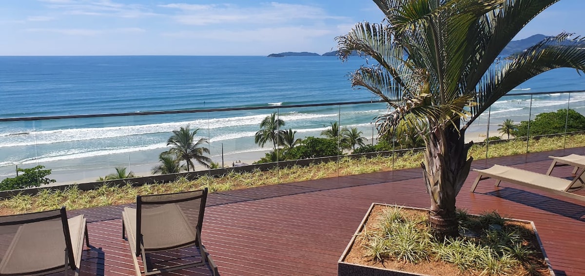 A spacious outdoor terrace offers a view of the ocean and coastline, with gentle waves lapping on the shore. Sun loungers are positioned nearby, surrounded by tropical plants. The area is designed for relaxation and appreciation of the scenic landscape.