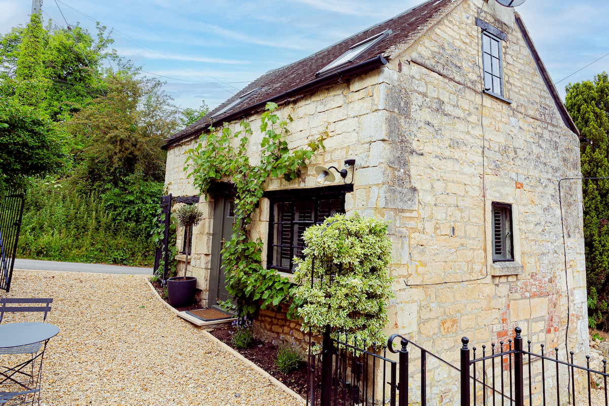 The cottage exterior showcases a traditional stone façade, framed by climbing greenery. Window shutters are closed, and a pebbled path leads to the entrance. A small seating area with a table and chairs is positioned nearby, inviting outdoor relaxation.