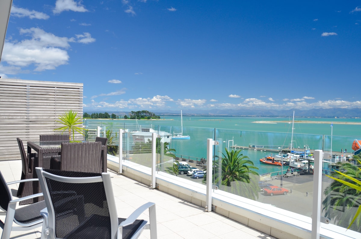 An outdoor terrace features a dining table surrounded by wicker chairs, overlooking the waterfront. A clear view of the water is visible, with boats anchored in the harbor and mountains in the distance under a bright blue sky.