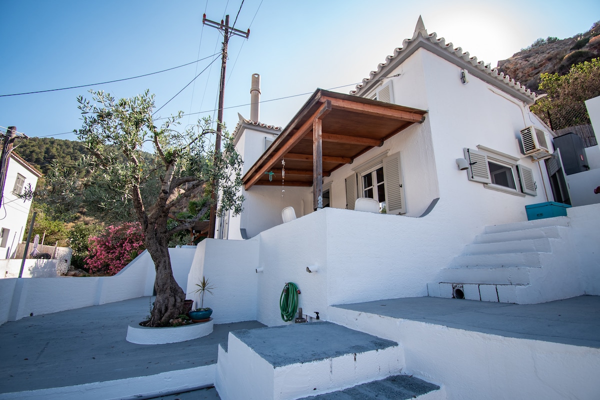 A white multi-level villa is showcased with a spacious patio area that features outdoor seating. An olive tree is positioned beside the entrance, providing a natural element. The structure is accented by lush green mountains in the background under a clear blue sky.