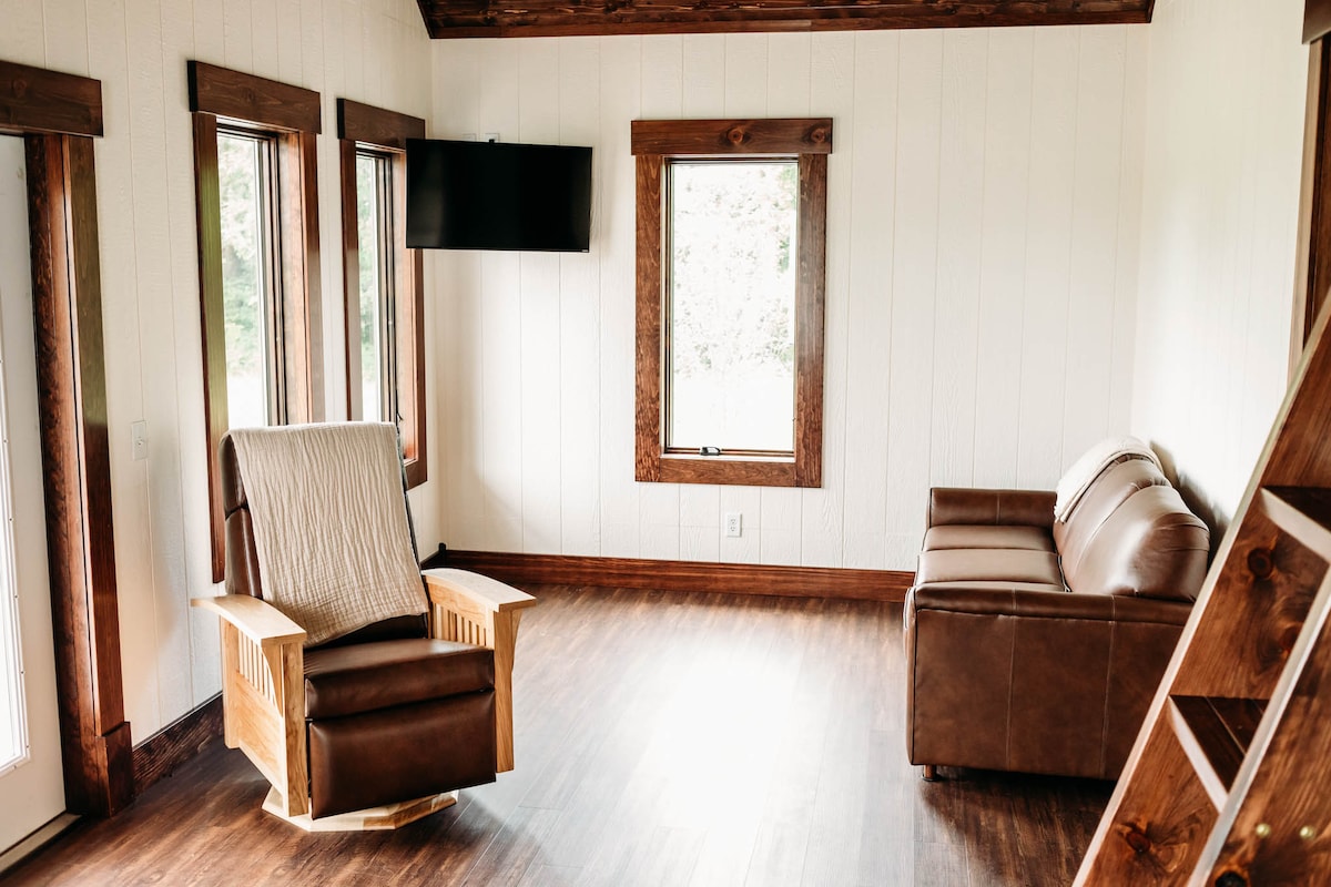 The living area features a recliner in warm wood tones beside a brown sofa. Natural light floods the space through large windows with wooden frames. A wall-mounted TV is positioned for viewing, and the flooring is a rich brown, enhancing the home's cozy atmosphere.
