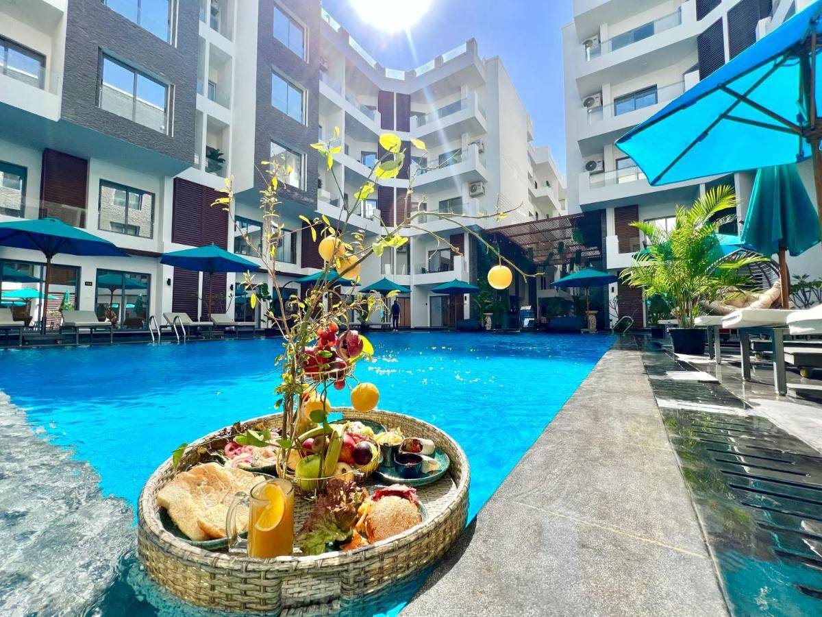 A tranquil pool area is surrounded by modern architecture, featuring multiple lounge chairs and umbrellas. A floating tray holds an array of fruits and beverages, adding a touch of color to the clear blue water. Lush greenery accents the inviting landscape.