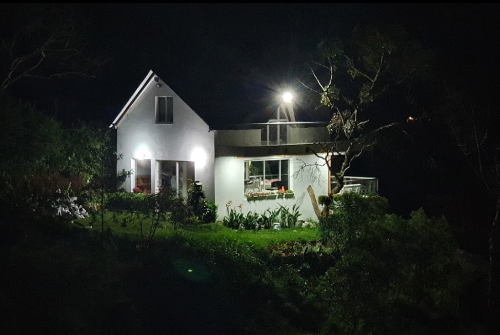 The house is showcased at night, illuminated by exterior lights. The two-story structure features large windows that provide glimpses of the interior glow. Well-maintained gardens surround the home, enhancing the serene atmosphere of the countryside setting.