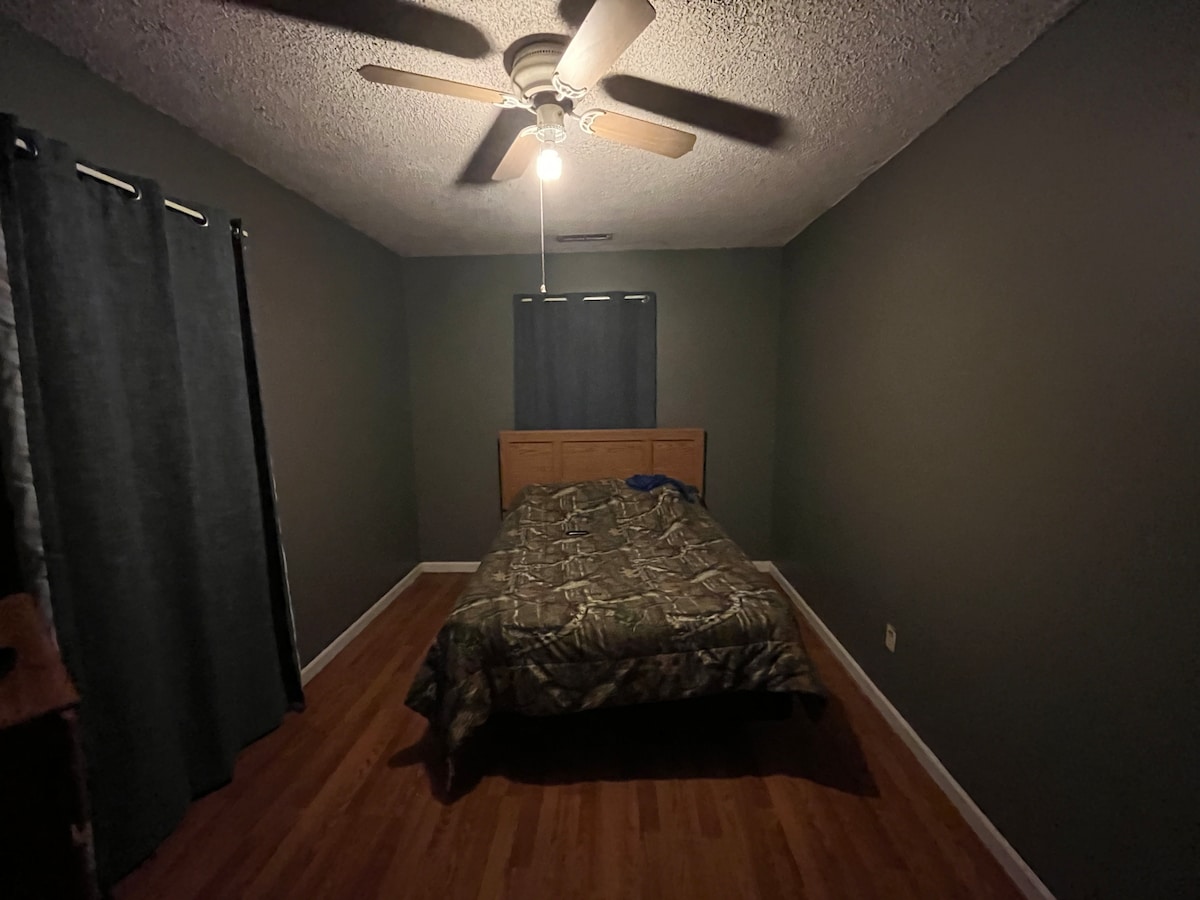 A simple bedroom is furnished with a spacious bed draped in a camouflaged comforter. Dark curtains cover the window, and a ceiling fan gently rotates above. Wooden flooring complements the earthy tones of the walls, creating a calm and restful environment.