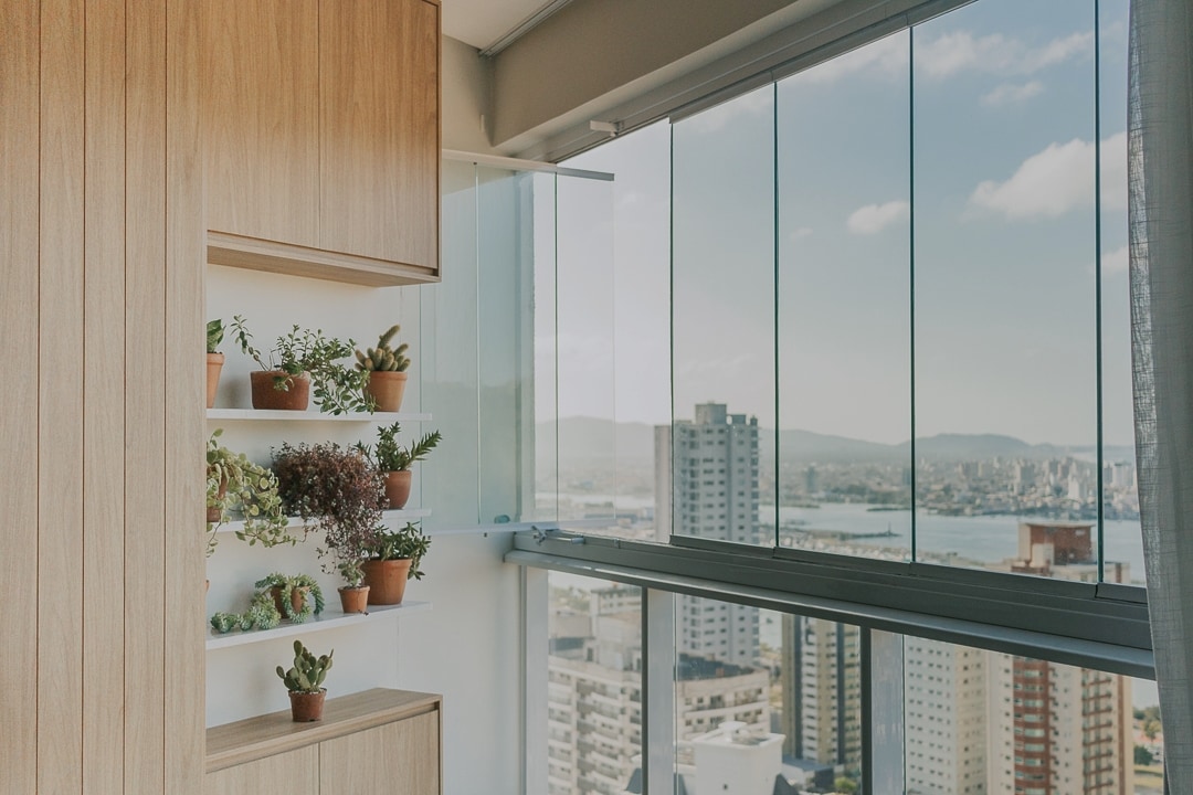 A light and airy space features a wooden shelf displaying a variety of potted plants. The floor-to-ceiling windows offer a sweeping view of the cityscape, mountains, and water, inviting natural light into the area.