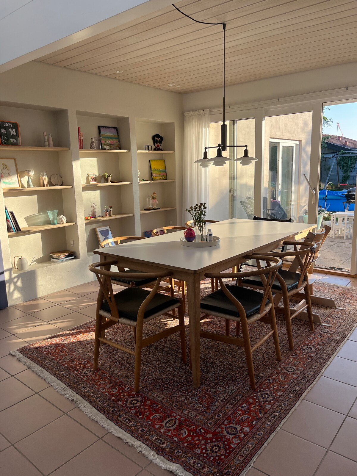 A dining area features a large white table surrounded by six wooden chairs with black cushions. Natural light floods the space through sliding glass doors, enhancing the warm tones of the room. Stylish shelving is displayed against the wall, showcasing various decorative items.
