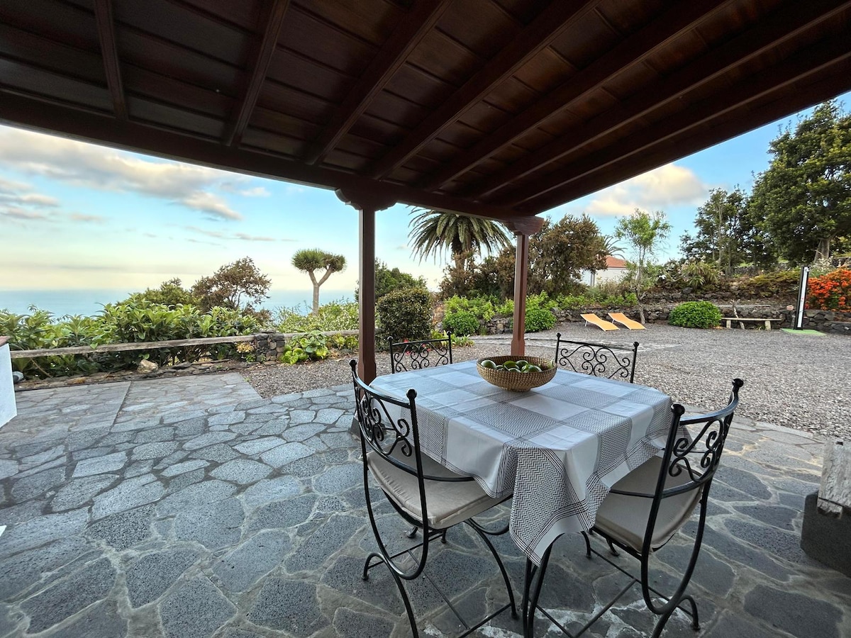 An outdoor dining area features a stone floor under a wooden canopy, with a round table set for six. Surrounding greenery and distant views of the sea can be seen, creating a serene atmosphere. Two sun loungers are positioned nearby for relaxation.