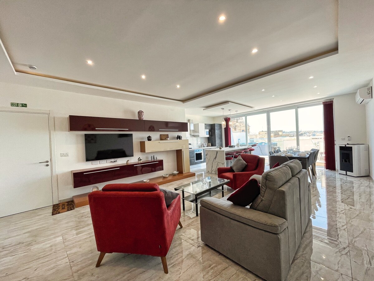 A spacious living area features modern furnishings, including a combination of red and grey seating. A sleek television is mounted on the wall, and large windows provide abundant natural light and views of the outdoors. Elegant marble flooring adds to the overall contemporary design.