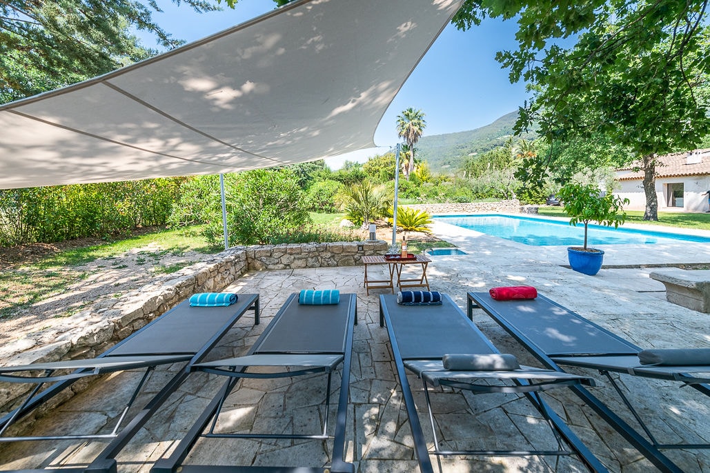 A spacious outdoor area features a set of four loungers arranged on a stone patio, each with colorful cushions. A large sunshade provides shelter from the sun, while a private pool sparkles in the background, surrounded by lush greenery and palm trees.