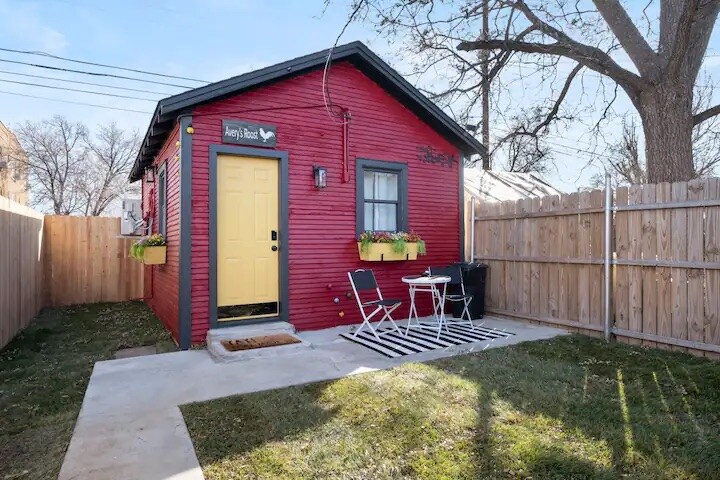A guesthouse exterior is featured, highlighted by a red facade and a bright yellow door. Potted plants adorn the windows, and a small patio with a striped rug and a table and chairs is visible. An enclosed yard is surrounded by a wooden fence.