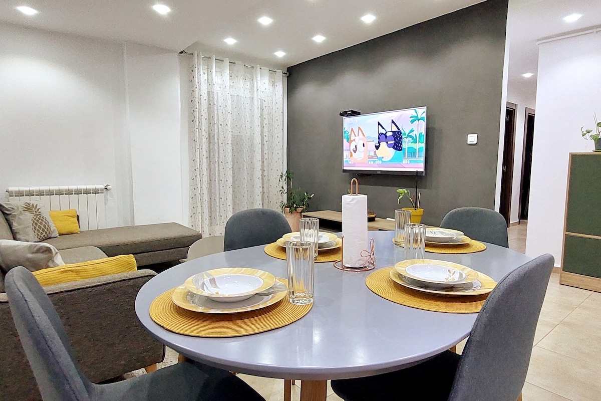 A modern dining area is set with a round table, adorned with plates, glasses, and woven placemats. A cozy sofa is positioned nearby, and a television is mounted on the wall, displaying colorful content. Soft lighting is provided by recessed ceiling lights.