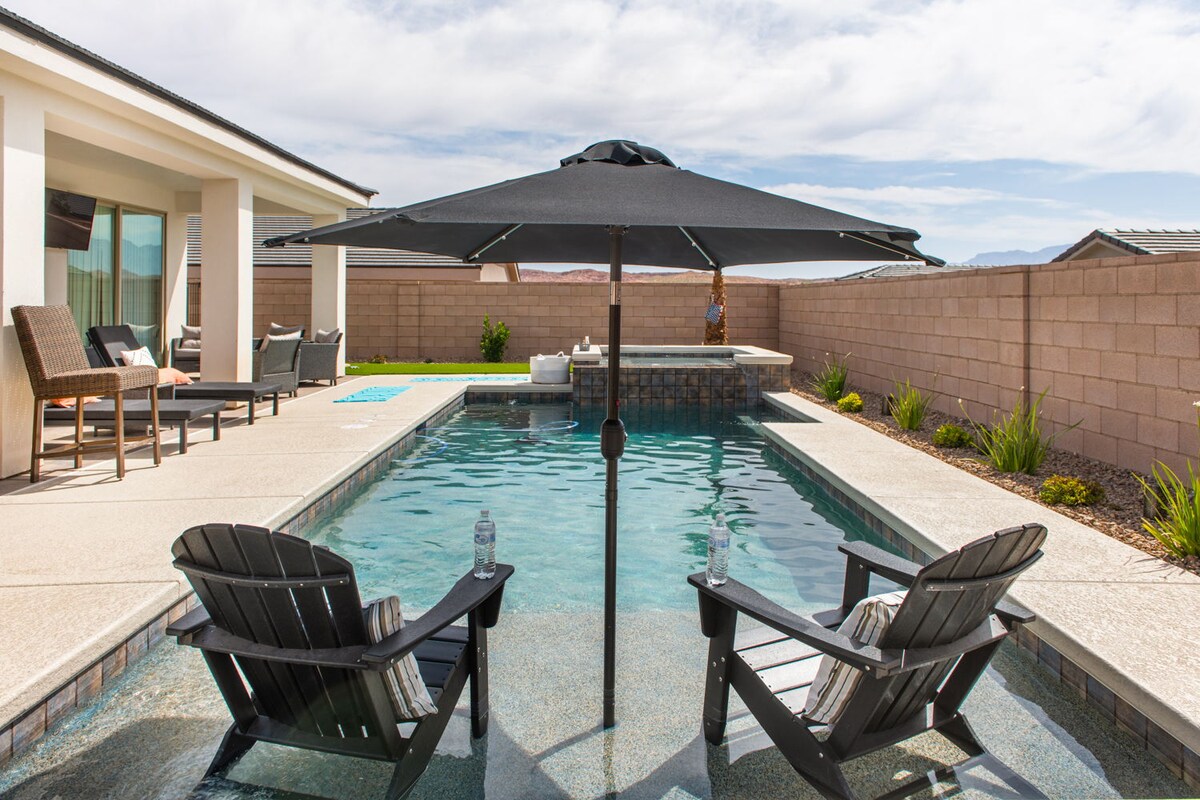 An outdoor space features a swimming pool with clear water, surrounded by a stone border. Two black Adirondack chairs are positioned under a large umbrella beside the pool, providing shaded seating. Lush greenery is seen along the pool's edge, enhancing the tranquil setting.