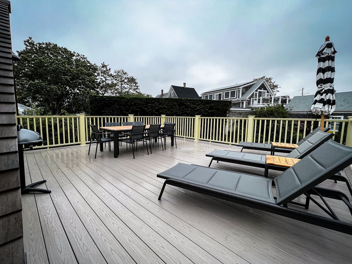A spacious outdoor area is shown, featuring a large dining table surrounded by chairs, alongside two lounge chairs. A striped umbrella provides shade, while a grill is positioned nearby. The railing offers privacy, and greenery is visible in the background.