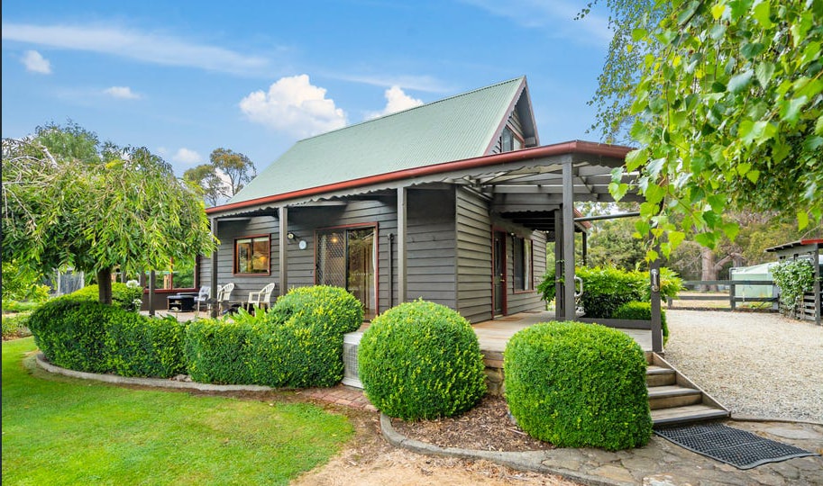 The exterior of a two-story shack is displayed, featuring a green roof and grey wooden cladding. Lush hedges surround the well-manicured yard, with a wooden deck offering seating. The tranquil setting is enhanced by trees and shrubs, providing a peaceful atmosphere.