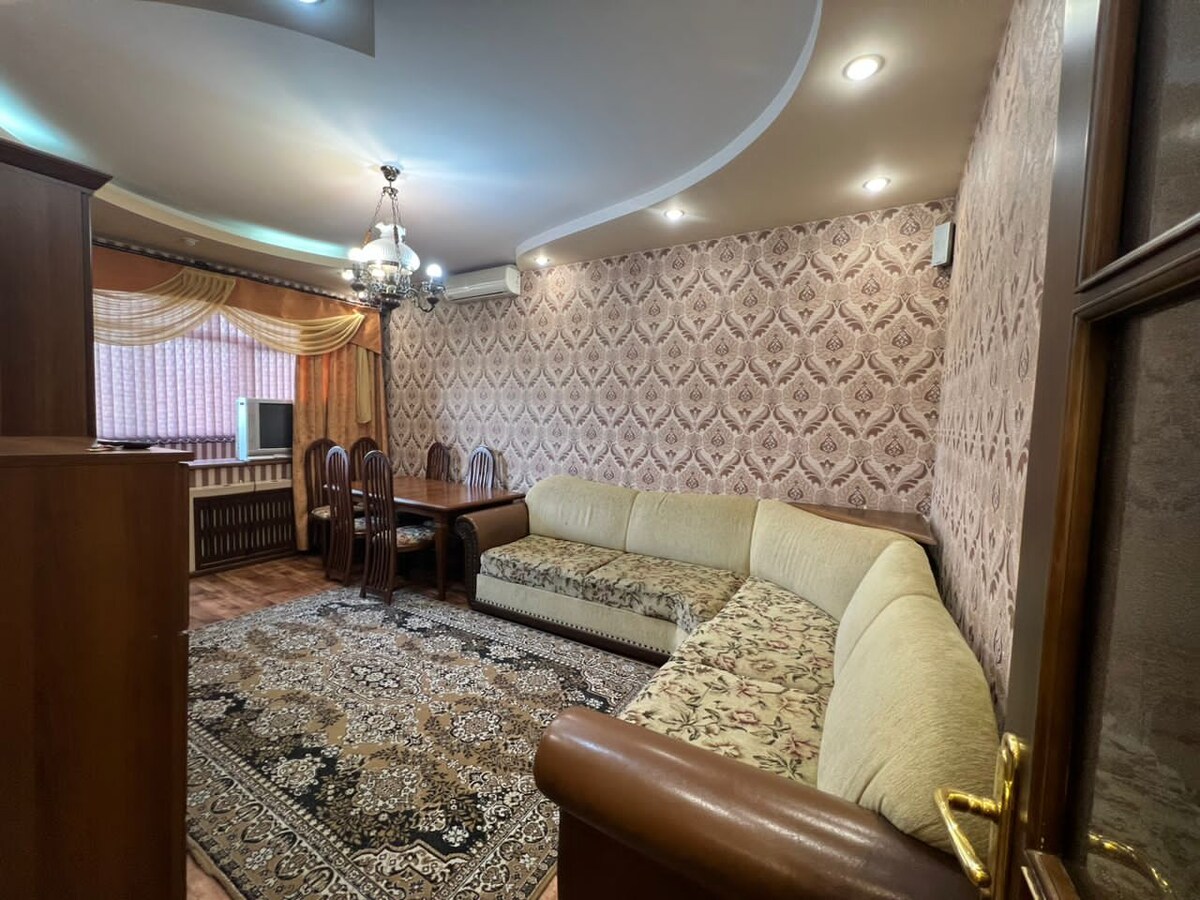 The living area features a spacious L-shaped sofa and a dining table surrounded by wooden chairs. Ornate wallpaper complements a patterned rug, while a chandelier hangs from the ceiling. Natural light filters through sheer curtains, creating a warm atmosphere.