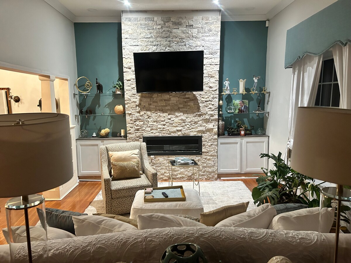 An inviting living room is accented by a stone feature wall and a mounted television. Comfortable seating is provided by a patterned chair and a spacious sofa. Light fixtures with soft shades and ornamental decor add subtle details to the warm ambiance.