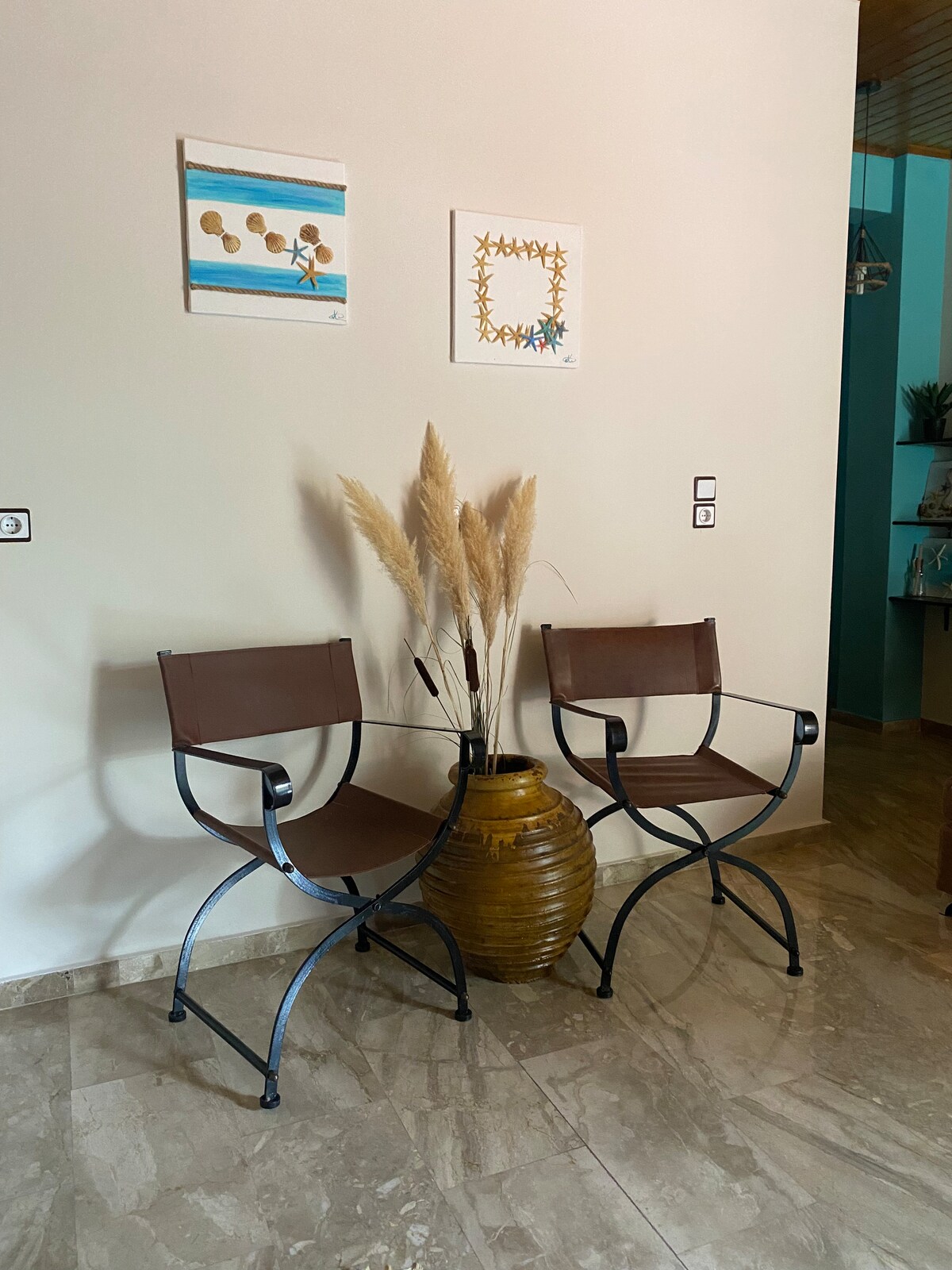 Two modern chairs are positioned beside a large terracotta vase filled with dried pampas grass. The neutral-colored wall is adorned with two pieces of framed wall art, adding a touch of creativity to the space. A marble floor enhances the overall aesthetic.