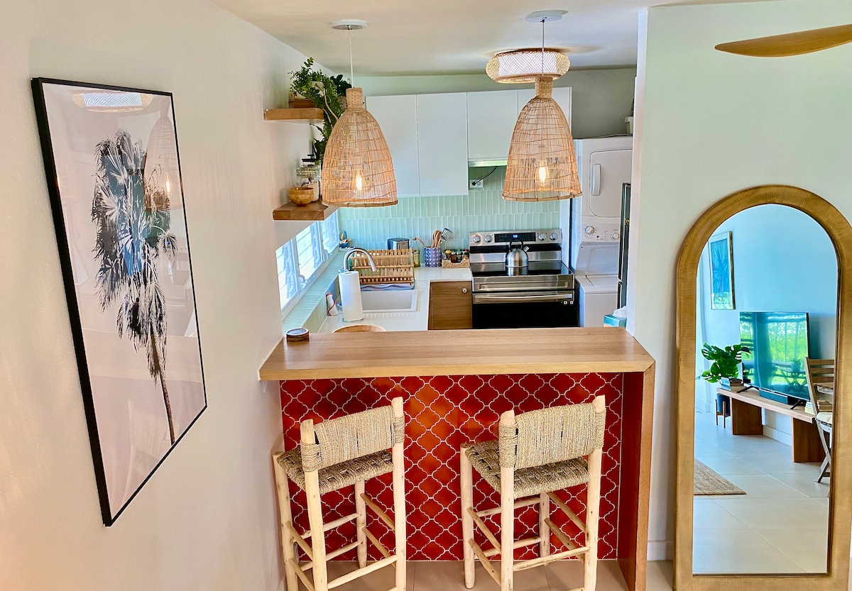 The modern kitchen features a spacious counter bar with two seating areas. Soft lighting is provided by pendant lamps above, highlighting the red tile wall. White cabinetry and appliances create a clean, bright atmosphere. A mirror reflects the inviting layout of the adjoining living area.