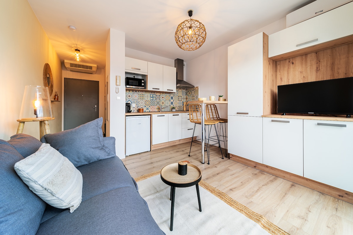 A modern studio apartment features a cozy sitting area with a blue sofa and a round coffee table. A well-equipped kitchen is visible, showcasing sleek white cabinetry and patterned tiles. A dining area with high-backed stools and a contemporary light fixture completes the inviting space.