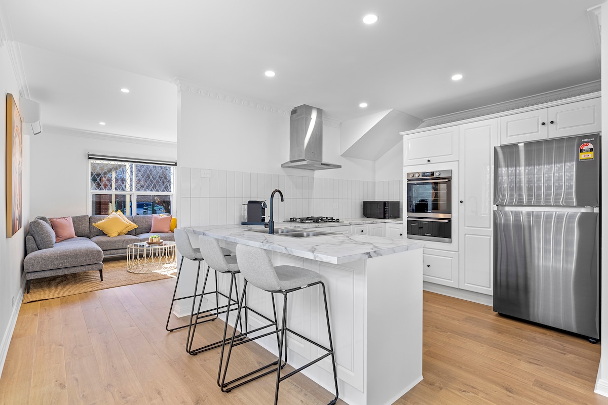 An open kitchen area features a marble island with three bar stools, offering a modern and welcoming space. Stainless steel appliances include a refrigerator and built-in oven, while natural light fills the room through a window, highlighting a cozy seating area with a sofa.