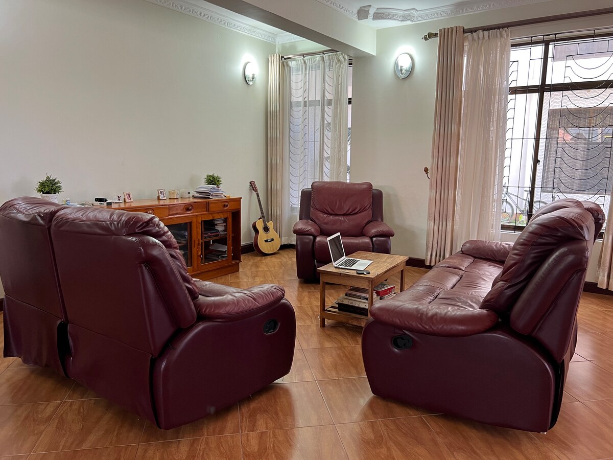 A spacious lounge area is showcased with two comfortable leather recliners arranged around a wooden coffee table. A guitar leans against the wall, while soft, natural light filters through sheer curtains covering large windows.