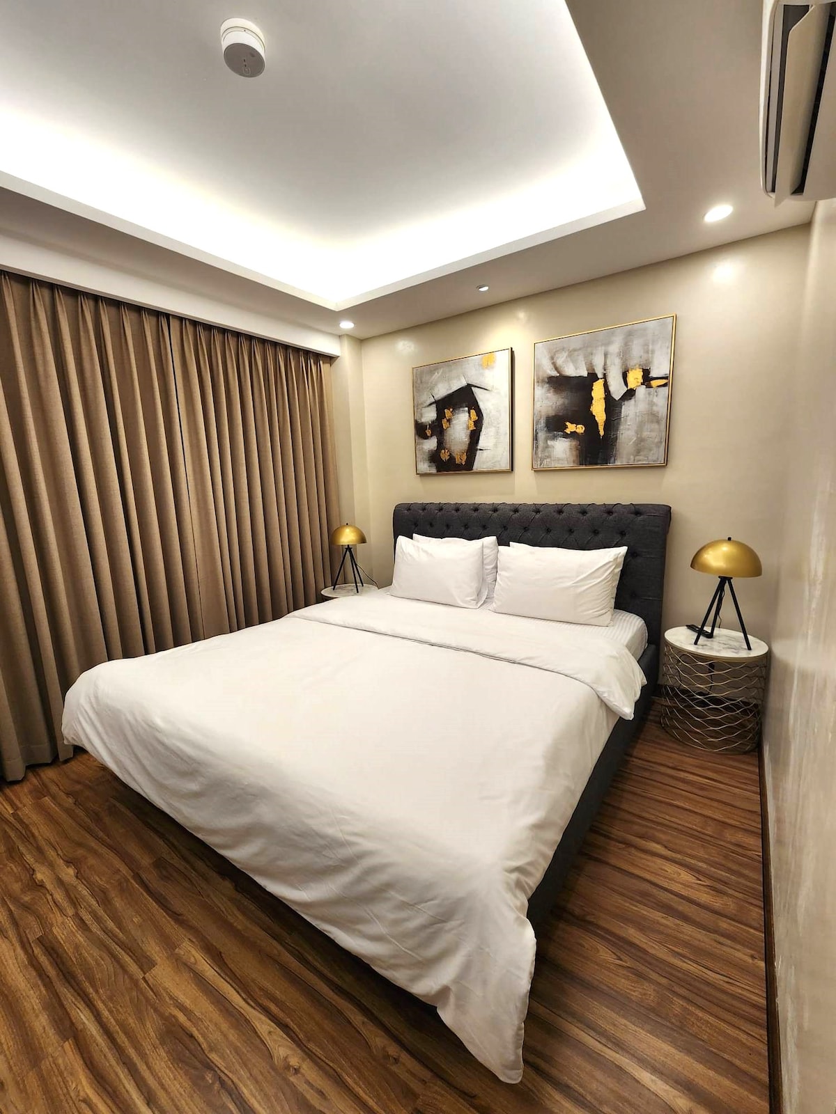 A well-appointed bedroom features a cozy bed dressed in white linens. Soft, warm lighting is provided by bedside lamps on modern side tables. The space is complemented by flowing drapes that frame the window and artwork adorning the walls, creating a serene atmosphere.