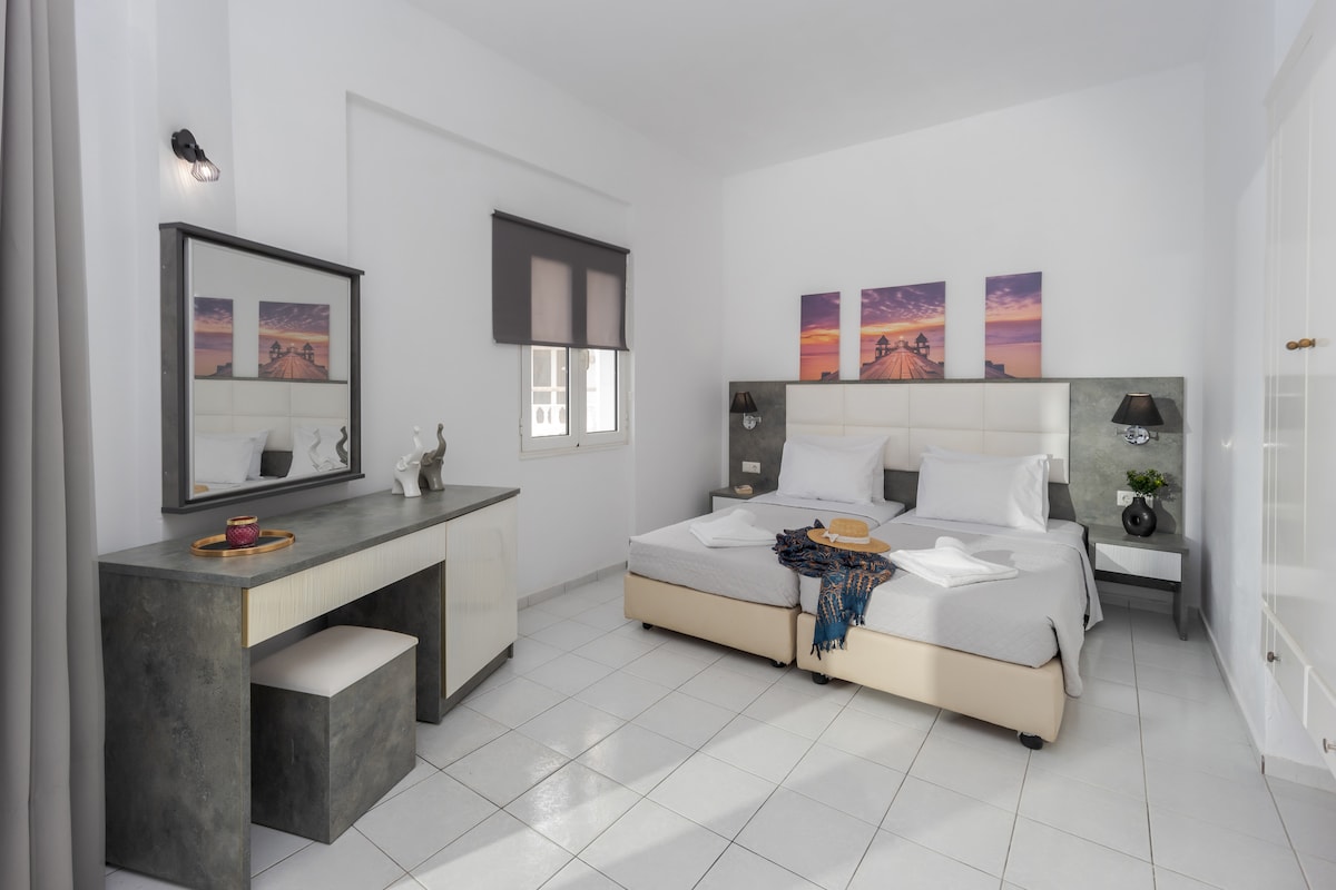A well-lit bedroom features a spacious bed with neutral bedding and a decorative throw. A modern desk with a mirror is positioned beside the bed, while artwork depicting a sunset adorns the wall above. Natural light enters through a window covered by light blinds.